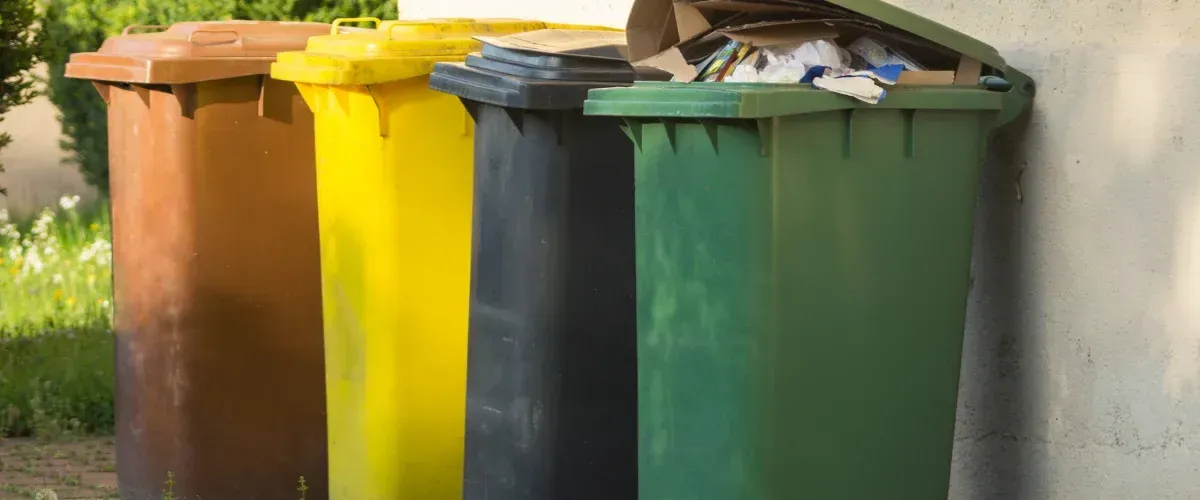 Four colorful recycling bins in brown, yellow, black, and green, full of waste, lined against a wall outside.