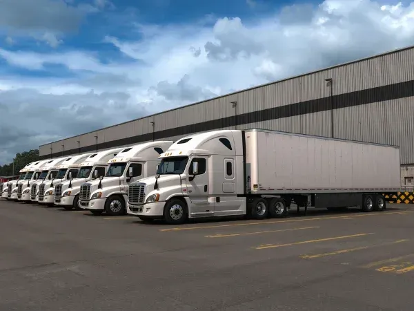 A long line of white semi-trucks with trailers parked neatly in front of a gray warehouse building under a partly cloudy sky.
