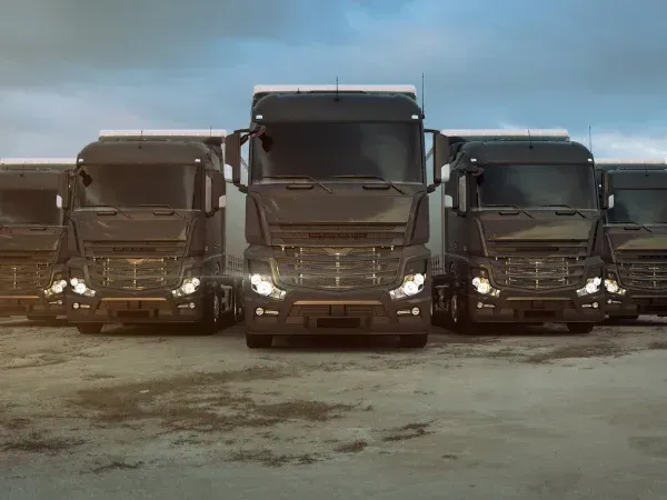 A fleet of dark semi-trucks with illuminated headlights parked in a row, facing forward under a cloudy sky.