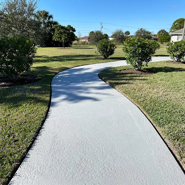 Finished concrete walkway curving through a backyard with simple landscaping on a sunny day.