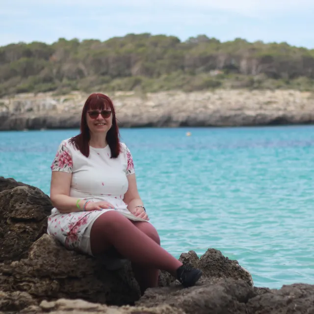 Frau mit Lipödem sitzt entspannt auf Felsen am Meer in Mallorca und lächelt in die Kamera.