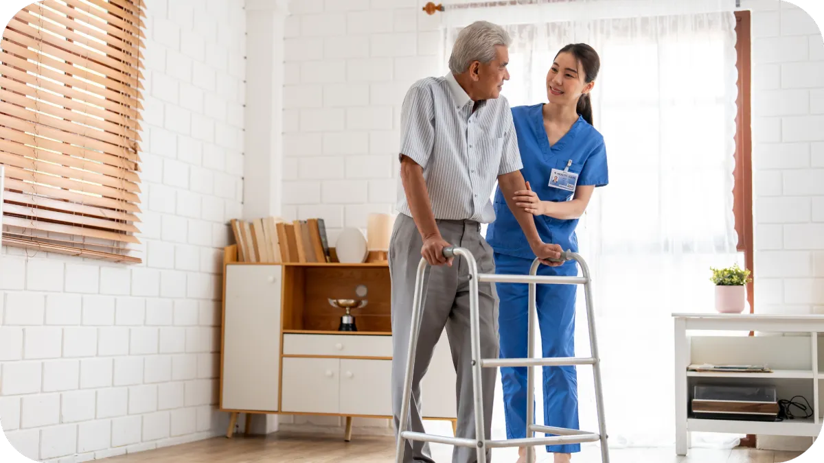 A female caregiver providing mobility assistance to a senior man using a walker in a home setting.