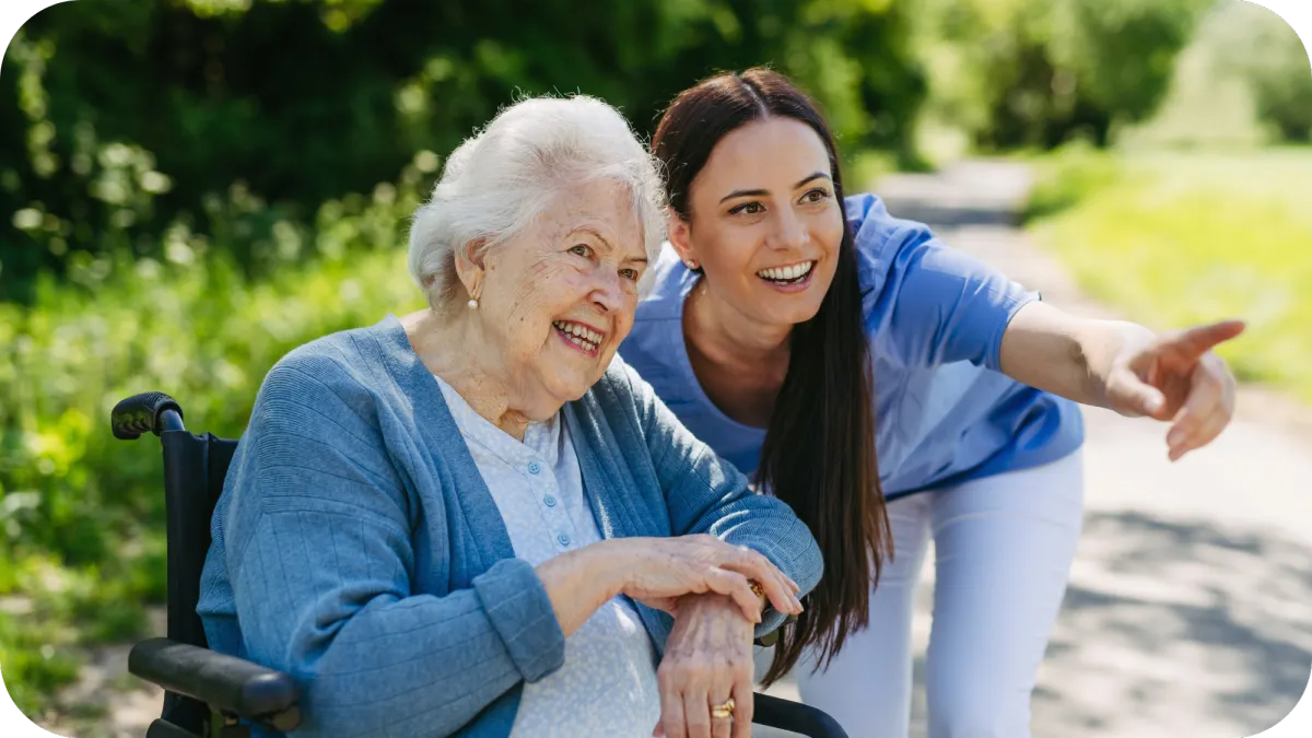 A caregiver and a happy elderly woman in a wheelchair enjoying a walk outside, with the caregiver pointing and smiling.