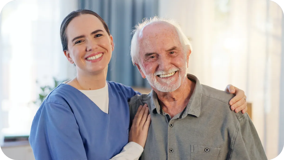 A smiling caregiver with her arm around an elderly man, both looking happily at the camera, representing a trusting home care relationship.