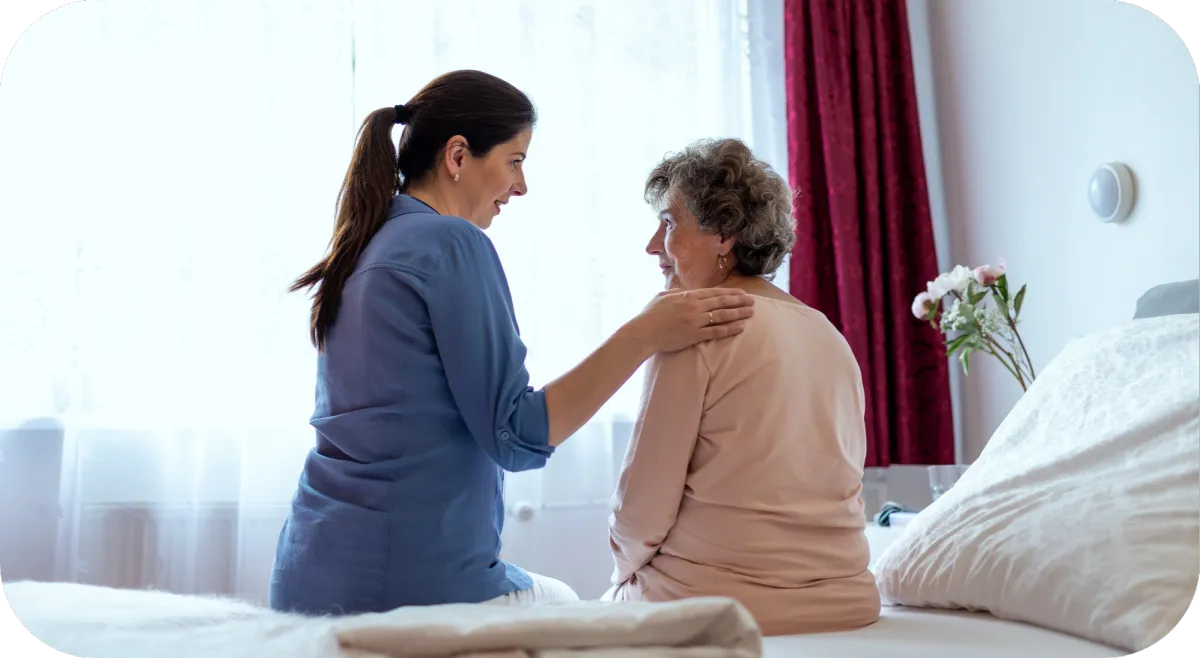 A caregiver sitting and talking with an elderly woman on a bed, offering comfort and emotional support.