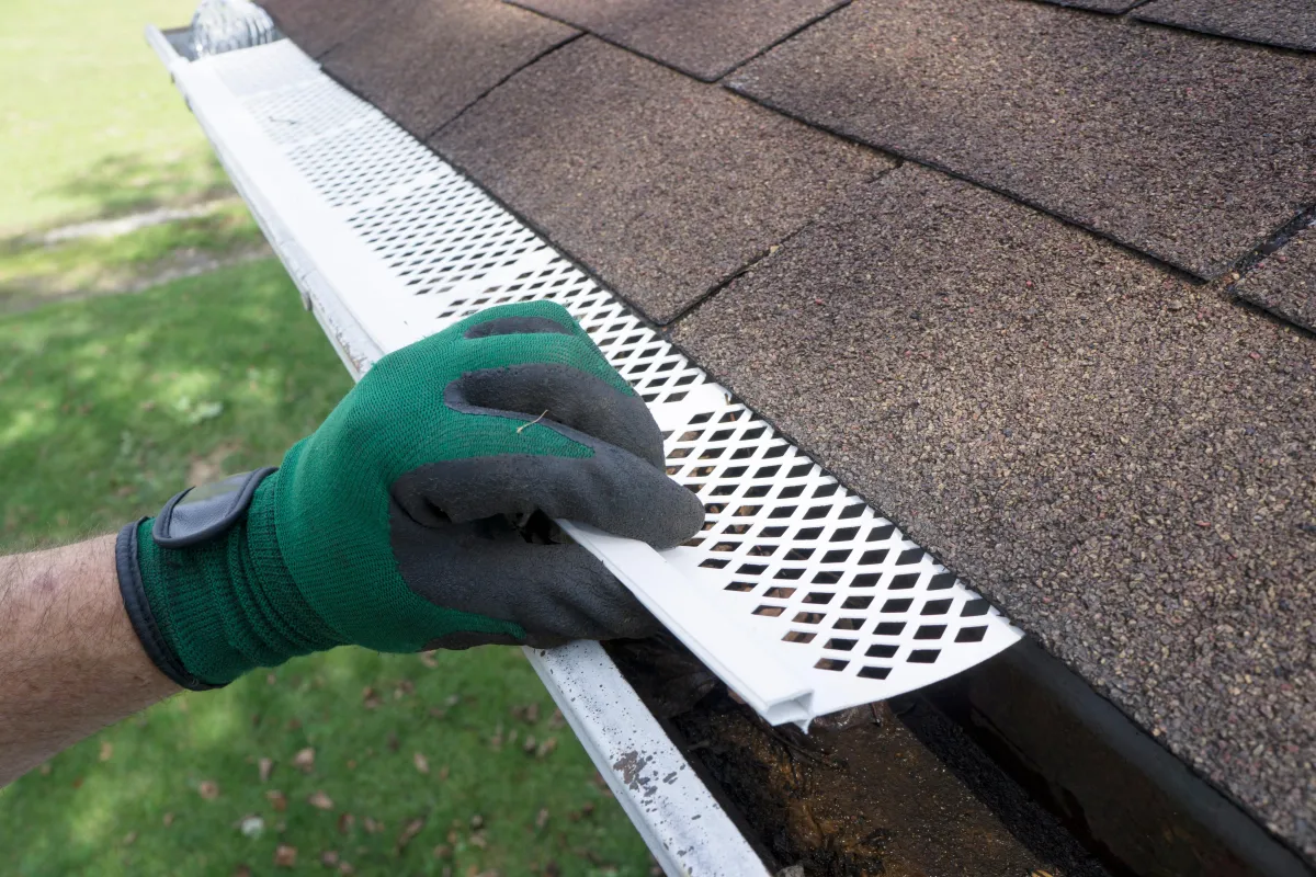 This image shows a man wearing green gloves installing plastic gutter guards below some 3 tab shingles on a sunny day.