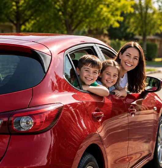 Smiling mother, son, and daughter looking out the windows of a red hatchback on a sunny suburban street.