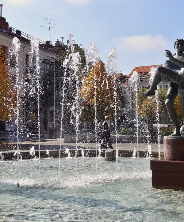 Public square fountain with statue and water jets surrounded by buildings.