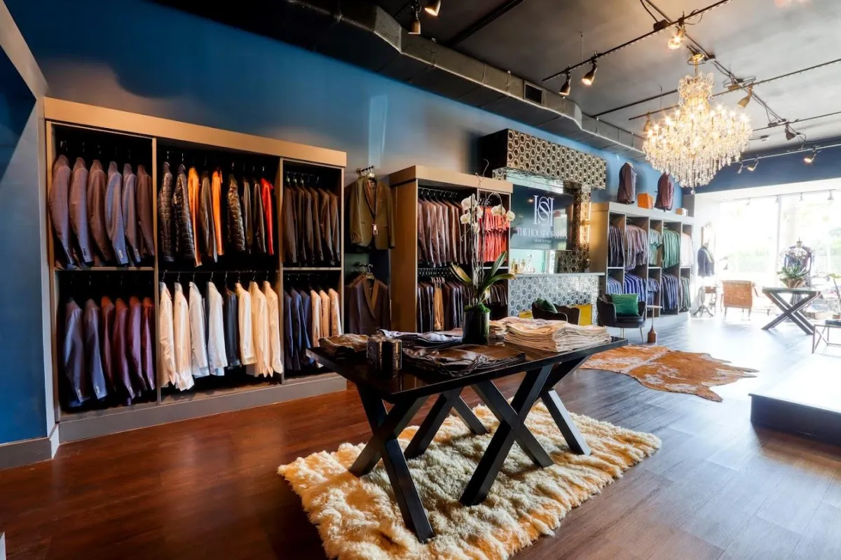 An elegant interior view of The House of Suits in Coral Gables, Florida, showcasing a sophisticated variety of premium suits on wooden hangers. Inviting, meticulously organized displays of complementary accessories effortlessly guide customers through the luxury shopping experience. The warm lighting and tasteful décor create a refined atmosphere that highlights the craftsmanship and attention to detail in every garment.