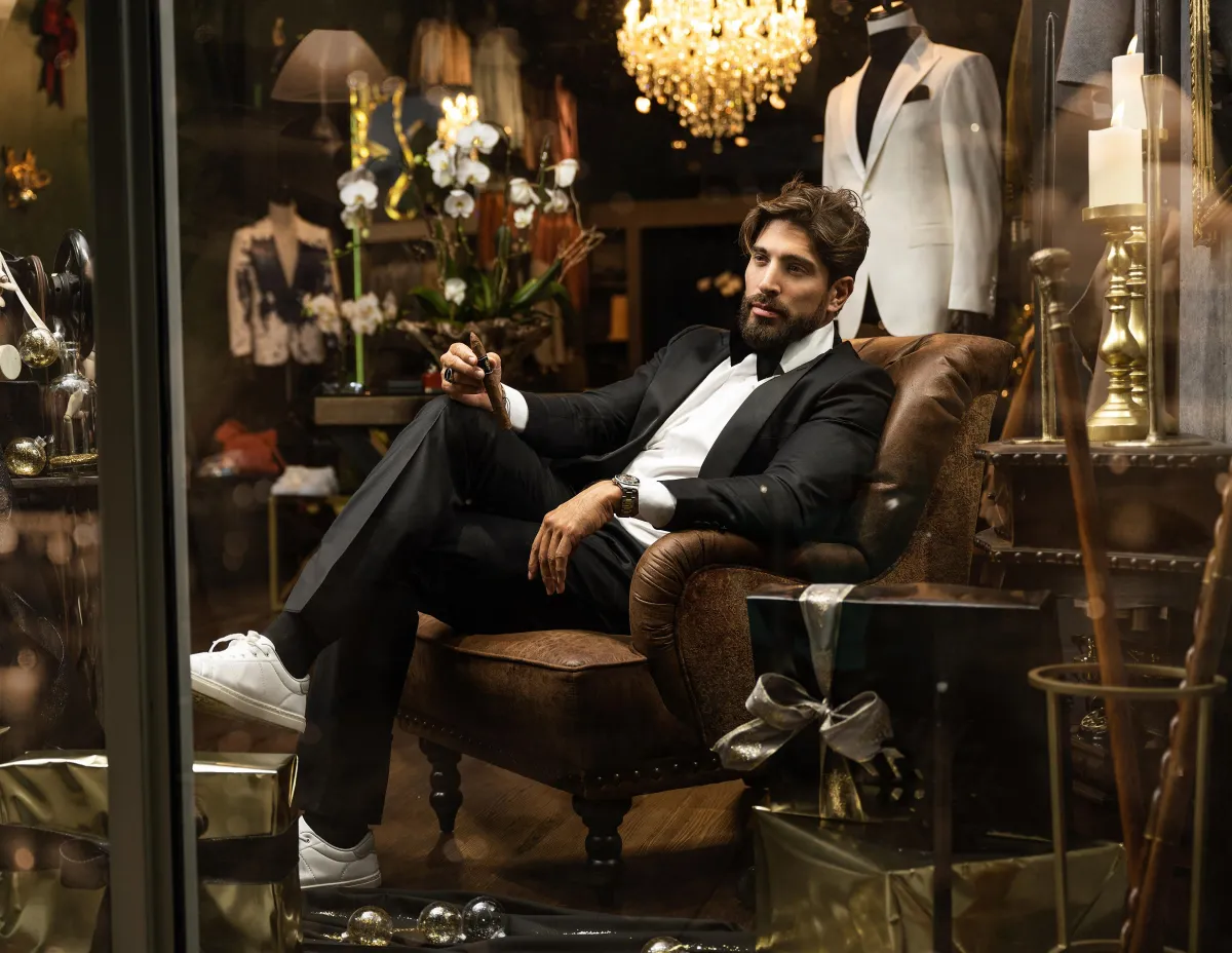 A sophisticated groom, wearing a perfectly tailored black suit, relaxes as he sits in a plush armchair, his fingers effortlessly holding an aromatic cigar. His intense gaze speaks of life's wisdom, as the dimly lit room highlights the elegance of his attire for the momentous occasion.