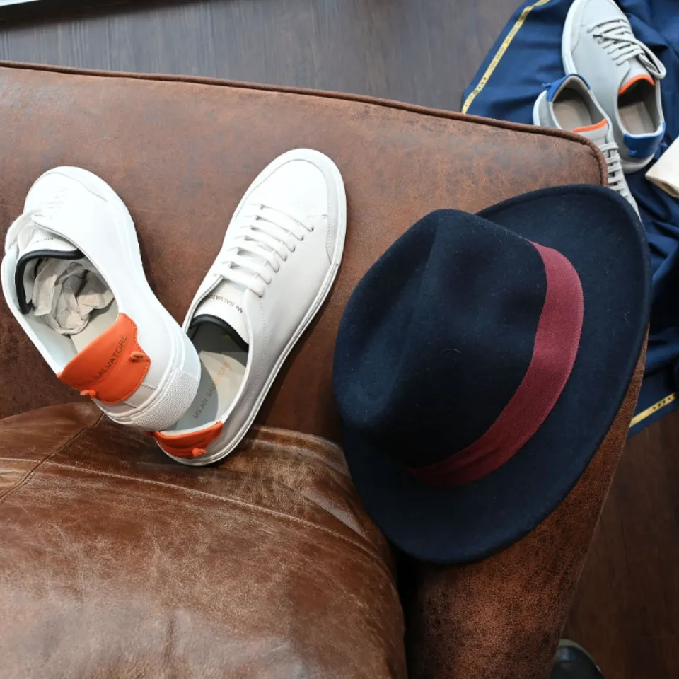 Image of a pair of stylish white casual shoes with rounded toes, comfortable rubber soles, and neatly stitched seams, complemented by an elegant black fedora hat featuring a classic dented crown, a slightly curved, wide brim, and a striking red grosgrain ribbon adding a tasteful pop of color.