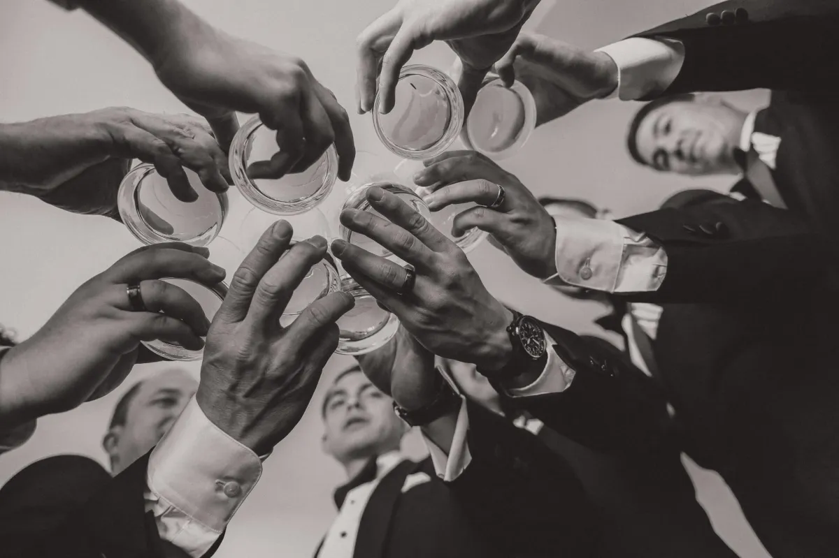 Sharply dressed groomsmen, donning impeccably tailored suits from "The House of Suit," raise a glass filled with champagne to toast. The grooms' suave attire exemplifies modern sophistication - all the groomsmen are wearing black suits with a white dress shirt.