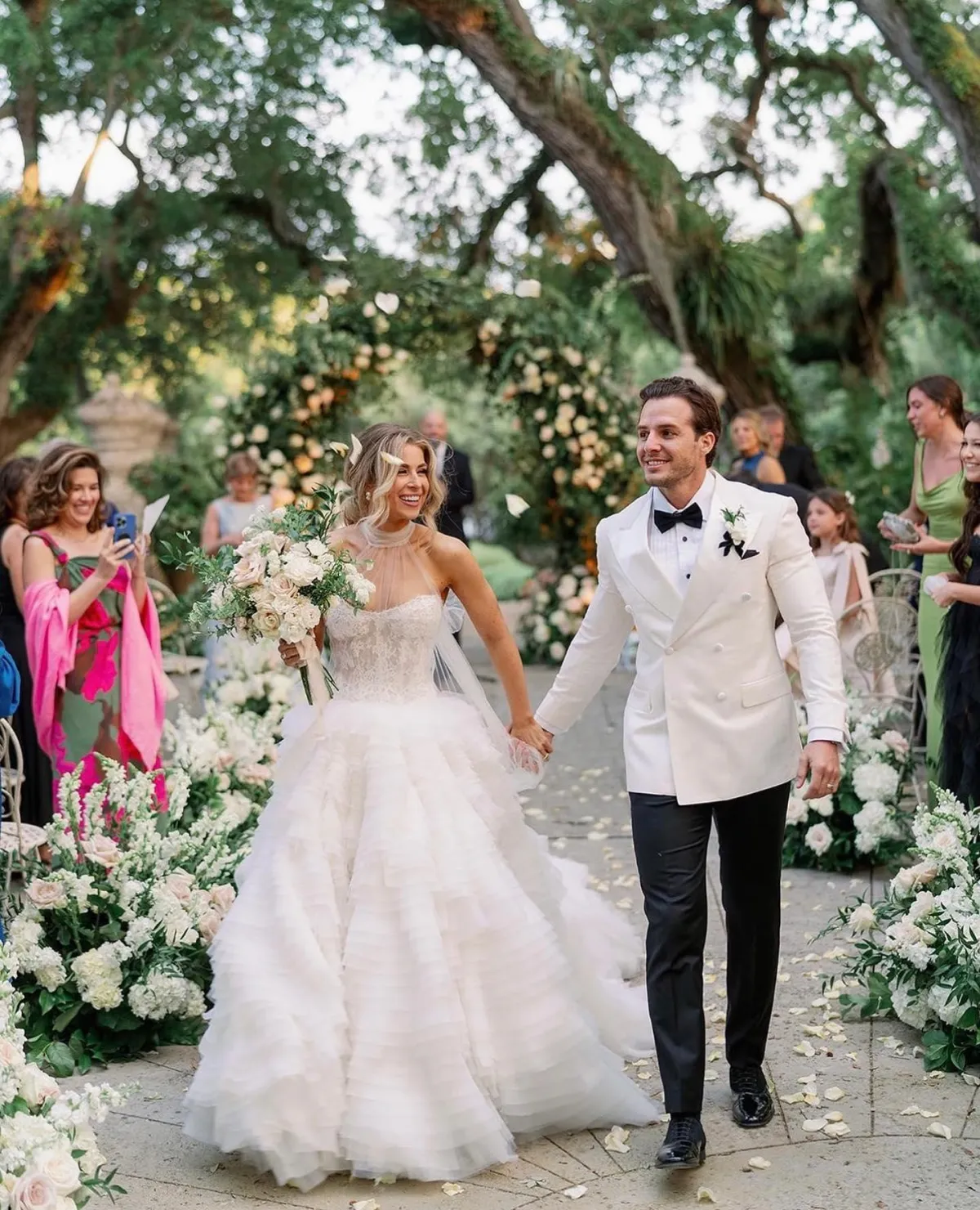 A dapper groom adorned in a tailored white tuxedo, crisp white dress shirt, and black bow tie, confidently standing next to a meticulously restored vintage car, its gleaming chrome accents and polished paintwork reflecting their timeless elegance as they both await the unforgettable ceremony.