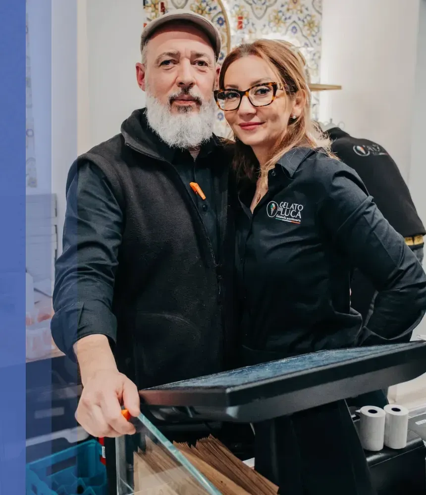 Founders of Gelato Di Luca standing inside their gelateria, wearing branded uniforms and smiling for the camera.