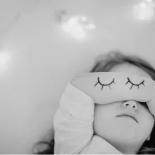 toddler sleeping in bed with arm over her eye, eyes drawn on her arm and fairy lights in the background