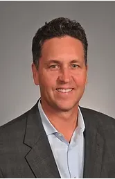 Portrait of a Black man in a tailored suit, standing in a bright boardroom, arms crossed, exuding leadership. The background shows a whiteboard with charts. 3:2 aspect ratio, sharp focus.