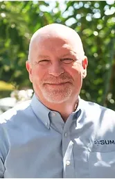 Portrait of an East Asian woman, seated at a conference table, reviewing documents, with a laptop open. The setting is a glass-walled office, 3:2 aspect ratio, natural daylight.