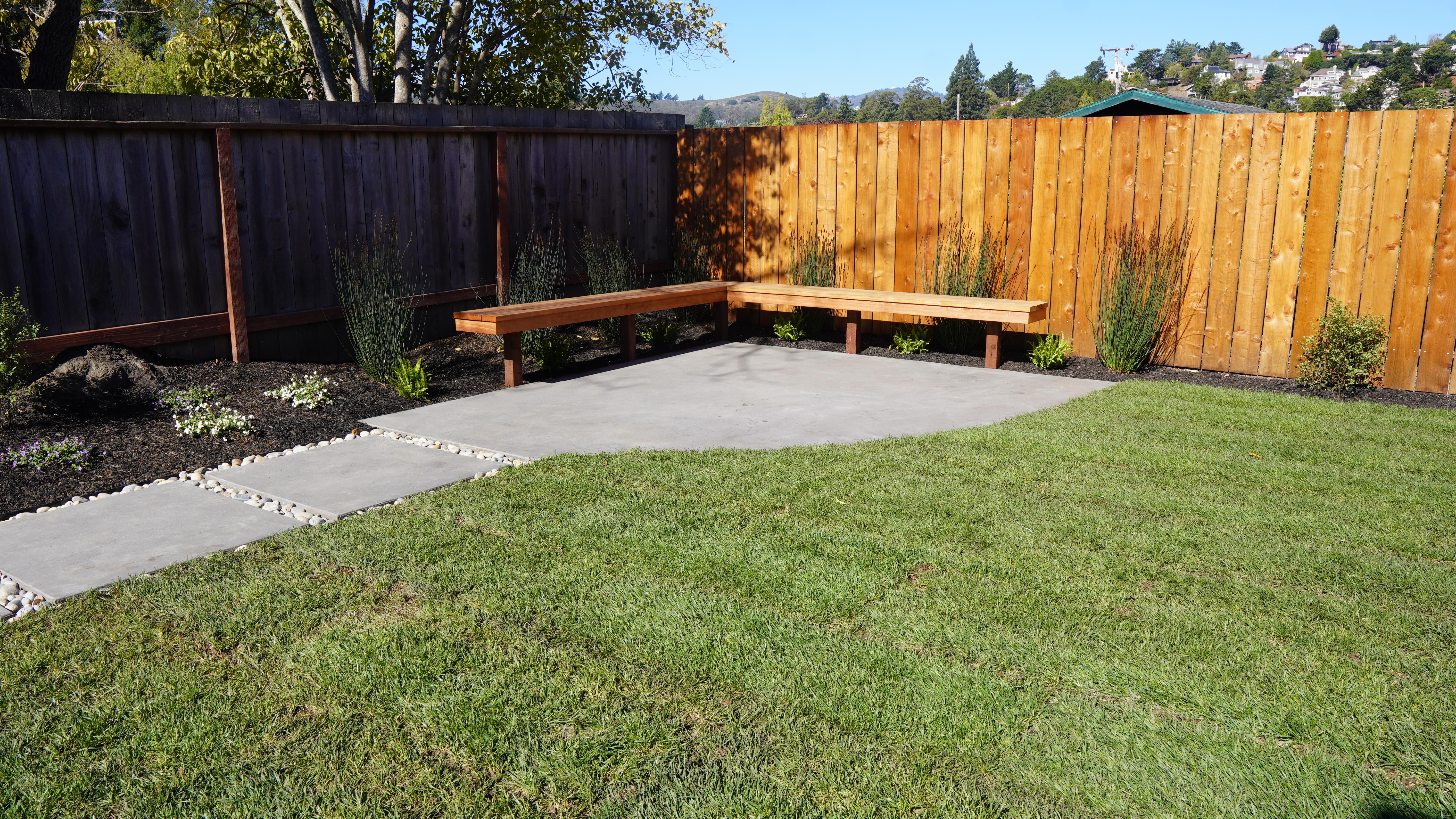 Back yard hardscape featuring new sod, concrete pathway and patio with gaps filled with pebble stones, wooden bench an plants behind the bench in Mill Valley, CA