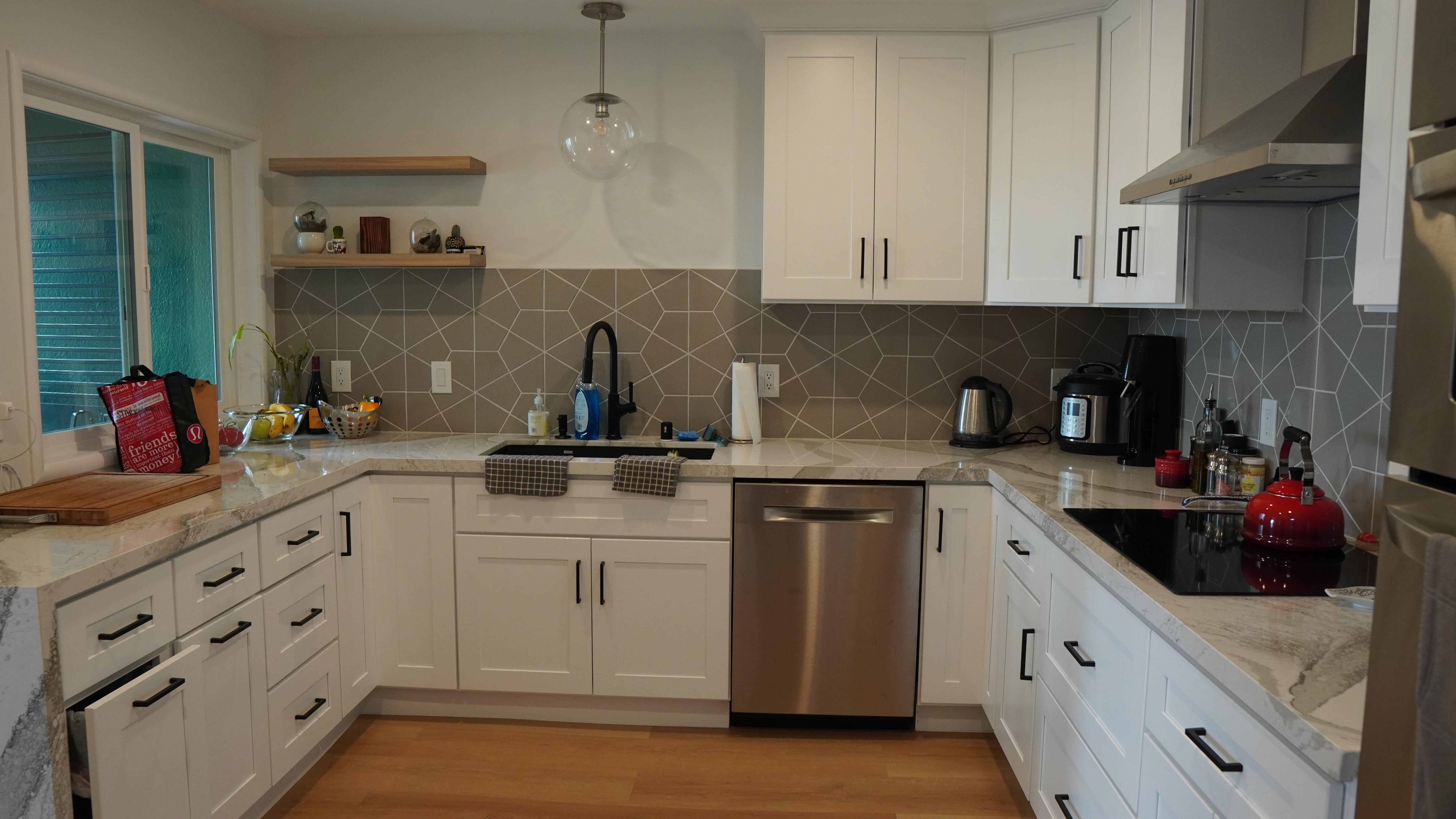 Kitchen remodel featuring white shaker cabinets with black hardware, grey backsplash tile, natural look floating shelves, and stain-less steel appliances in Daly City, CA.