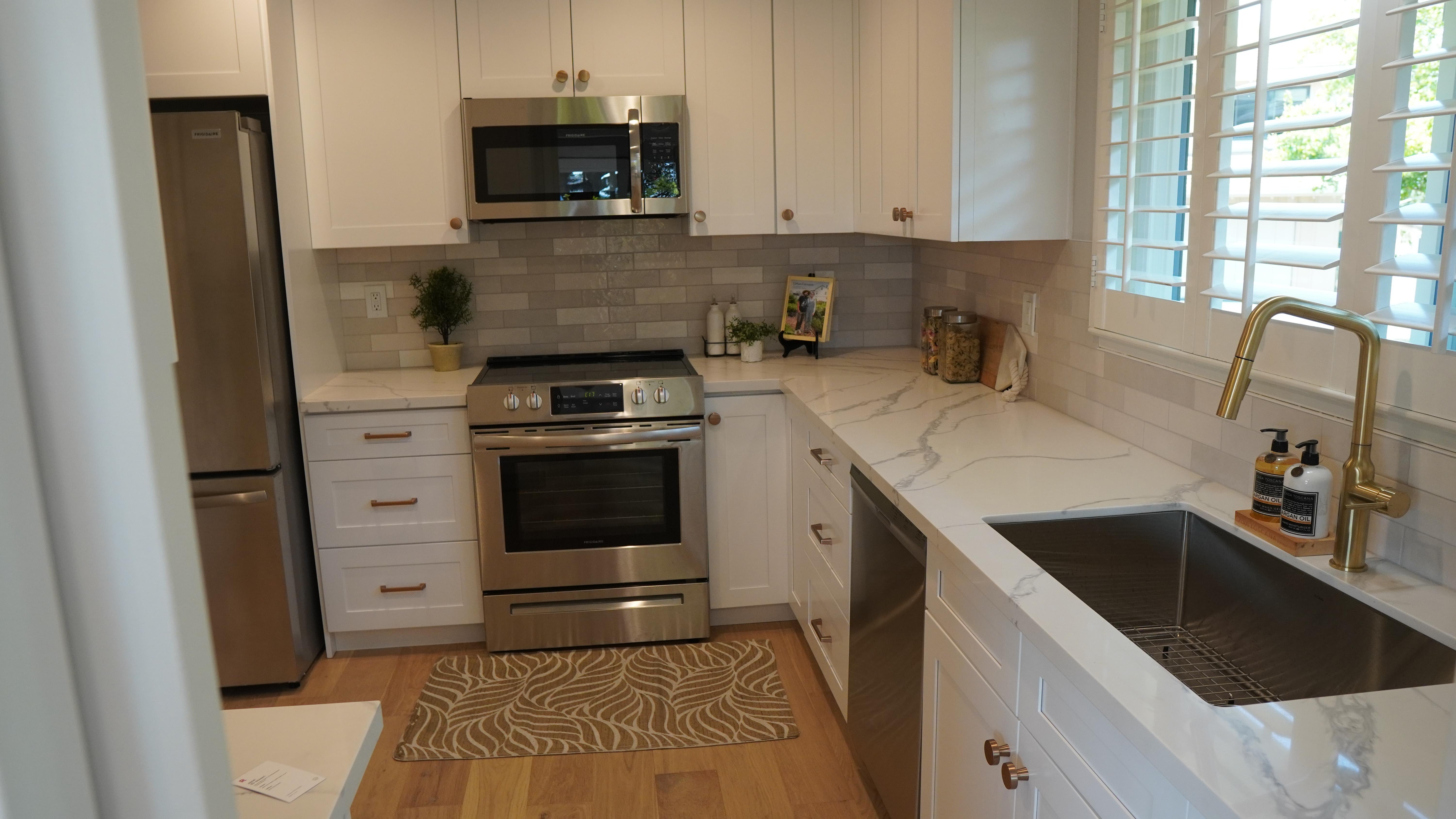 kitchen remodel featuring white cabinets with golden hardware, white calacata quart countertops, grey tile backsplash, stainless steel appliances, golden sink faucet, window with curtains in San Carlos CA.