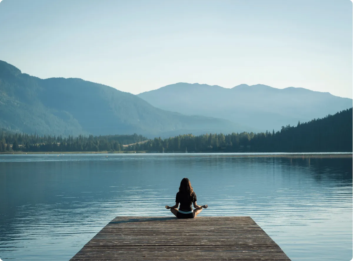 A person sits cross-legged at the end of a wooden dock facing a calm lake and layered mountains under a clear sky.