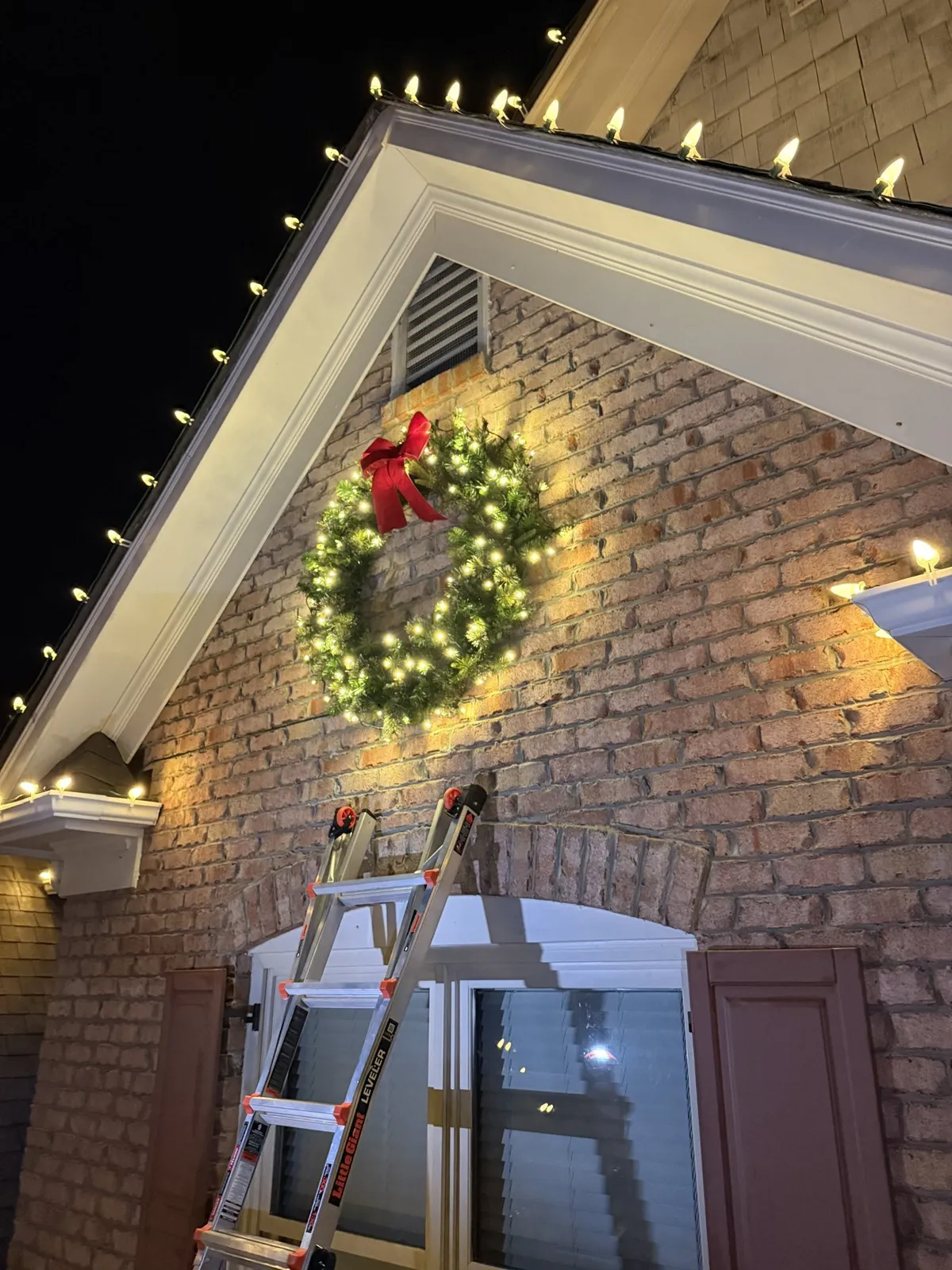 Lit wreath with a red bow on a brick gable, with warm white bulbs along the roofline