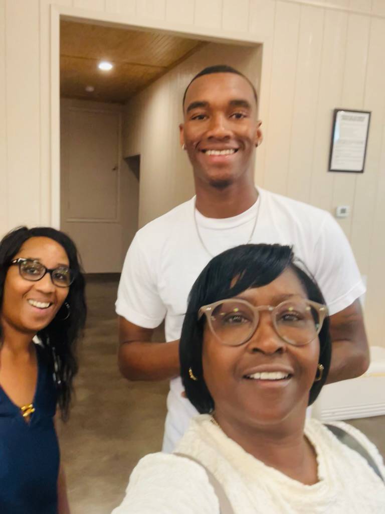 A vibrant group photo of extended family members of all ages gathered on a picnic blanket in a sunny park, everyone smiling and some waving at the camera, with trees and balloons in the background.