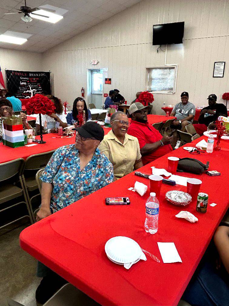 Two elderly relatives sitting on a bench, animatedly sharing stories and laughing, surrounded by younger family members listening intently, with a festive outdoor setting in the background.
