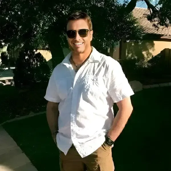 A man wearing a white short-sleeve shirt and sunglasses, known as Alexandar 1111 the astrologer, stands outdoors with hands in pants pockets, smiling.