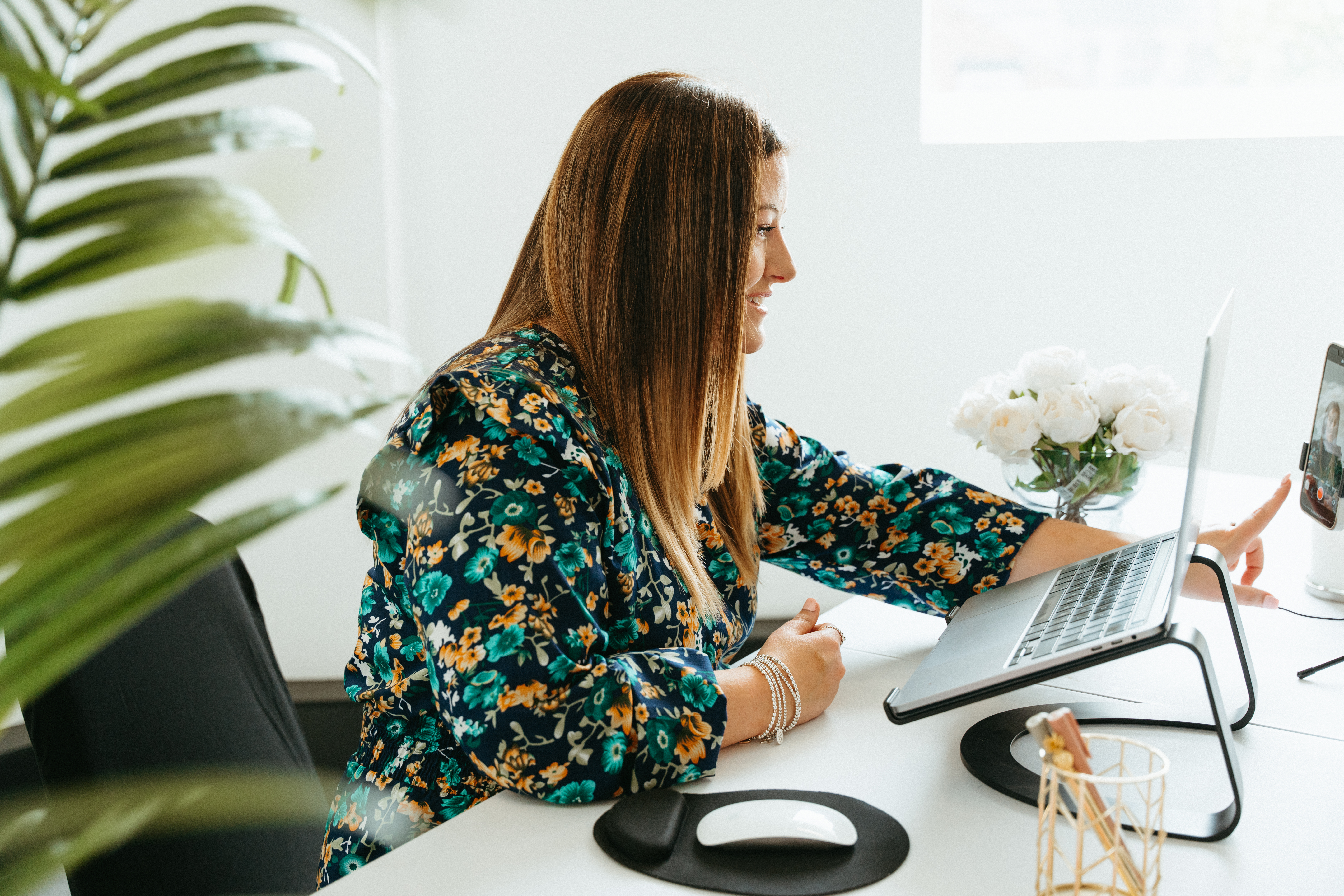 A laptop on a wooden table displays a Zoom meeting with a smiling female presenter and several participant video tiles. A notepad, pen, and a cup of tea are nearby. The room is softly lit, with a plant in the background, evoking a welcoming, organised home workspace.