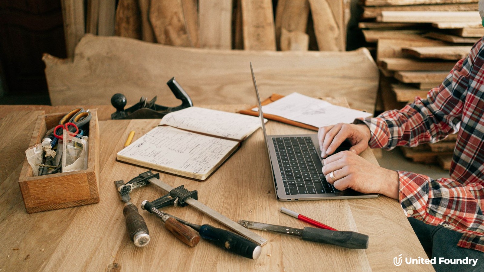 A tradesperson in a plaid shirt working on a laptop at a workbench with hand tools and a notebook