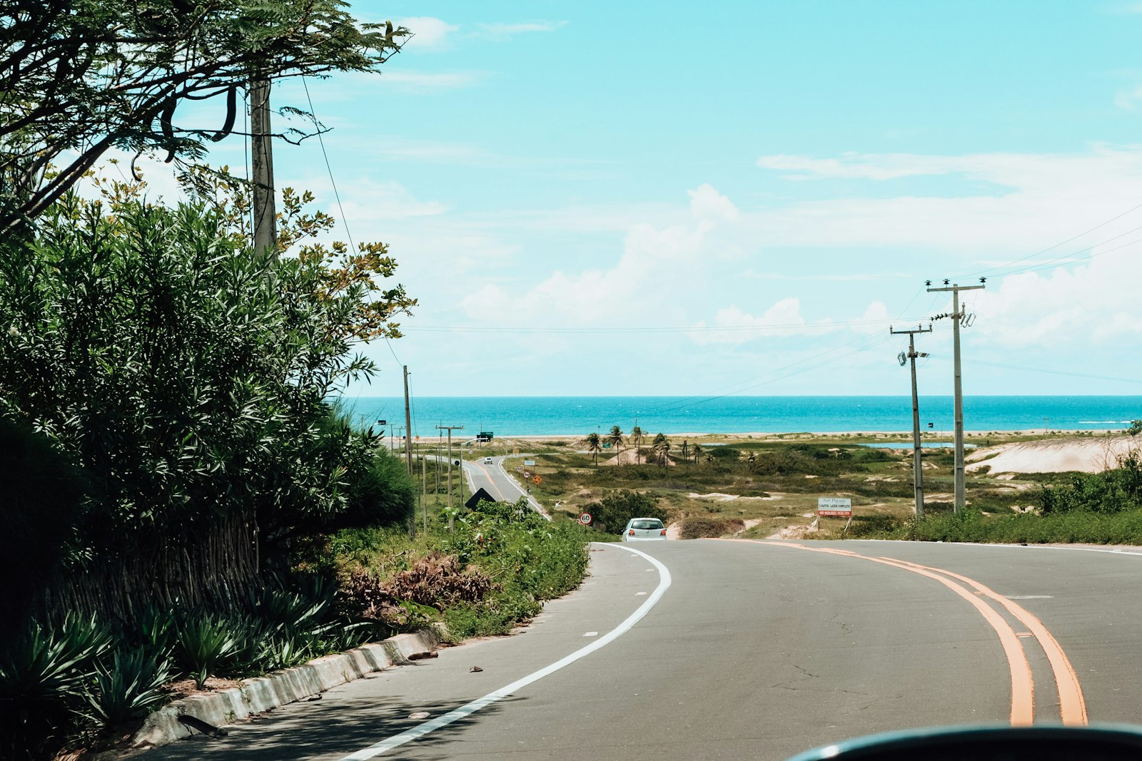 Coastal road curving toward the ocean under a bright sky, seen from behind the wheel on the drive to the beach