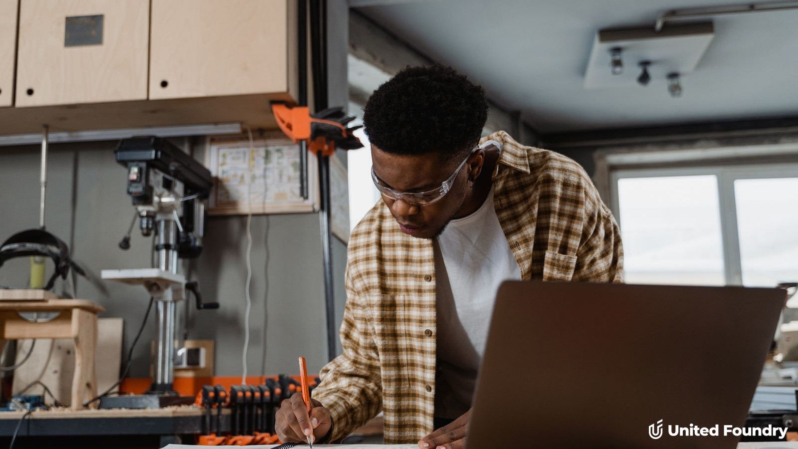 Contractor planning a project at a workshop bench with a laptop and notebook, United Foundry logo