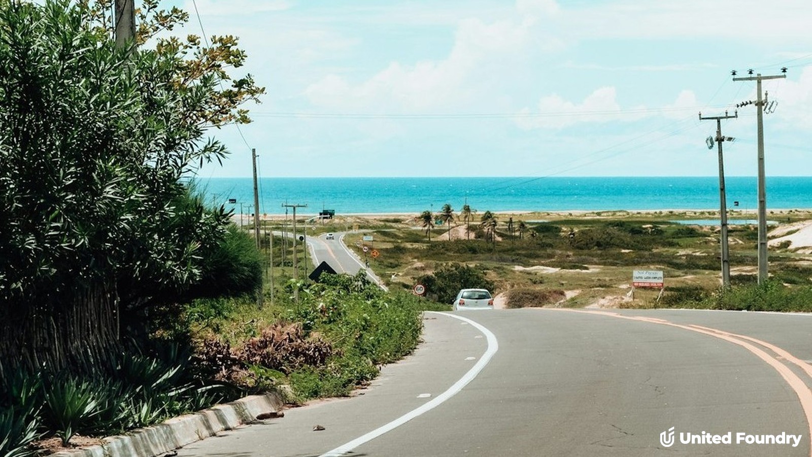 Coastal road curving toward the ocean under a bright sky, seen from behind the wheel on the drive to the beach