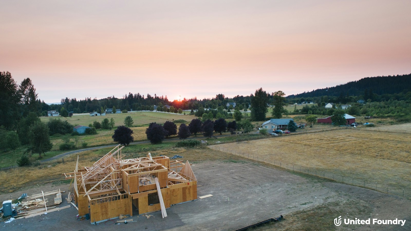 Aerial view of a custom home under construction with wood framing at sunset, surrounded by open fields