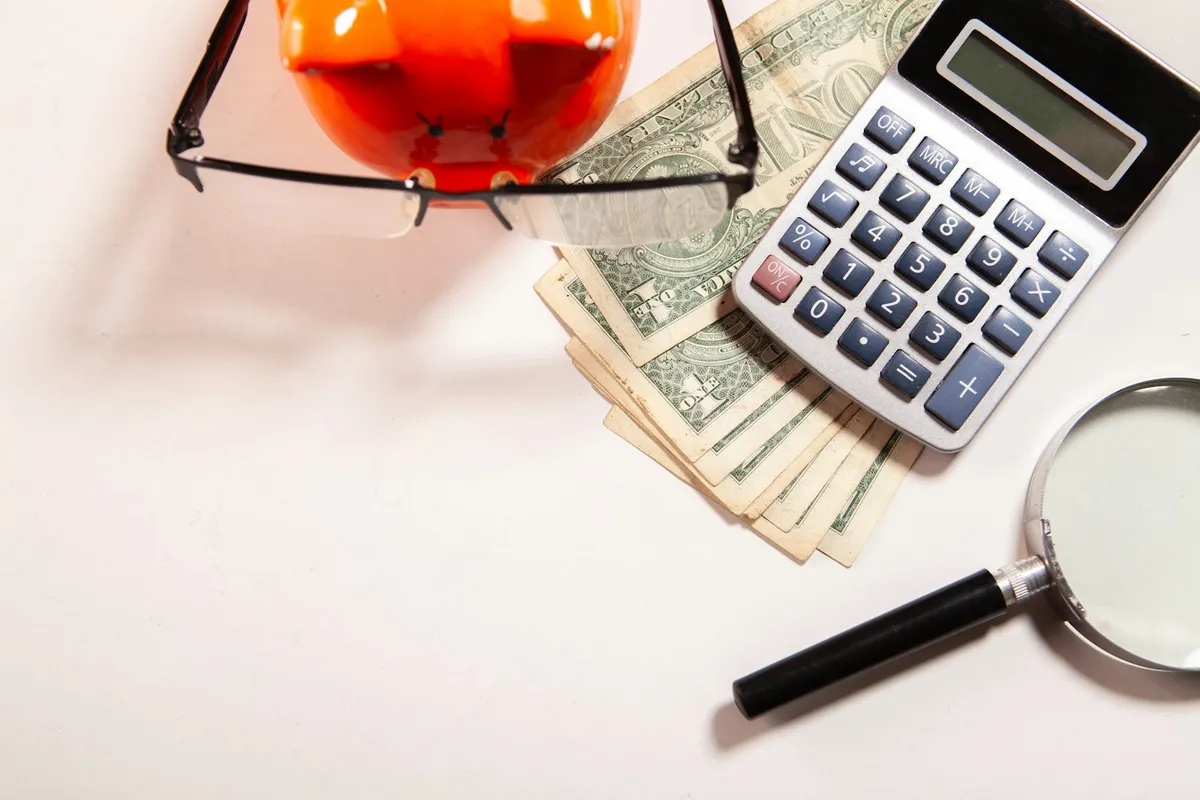 Calculator, piggy bank, cash, and magnifying glass laid out on a desk, representing budget analysis for Local Services Ads categories