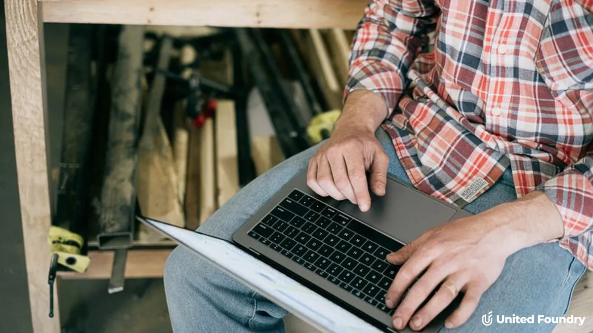 Tradesperson in a plaid shirt working on a laptop in a woodworking shop, United Foundry branding