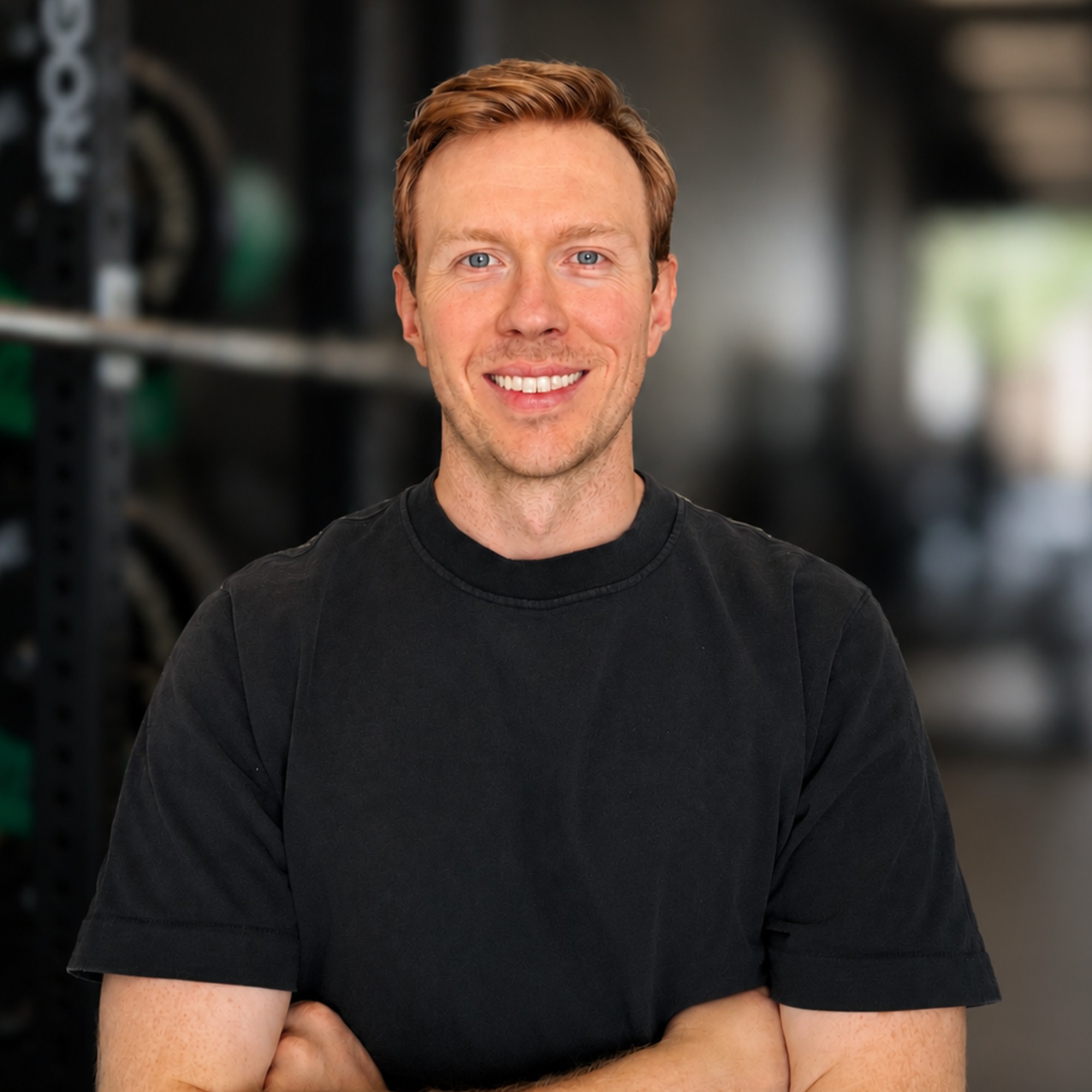 Portrait of a smiling man with short reddish-brown hair wearing a plain black t-shirt, arms crossed, standing in what appears to be a gym.