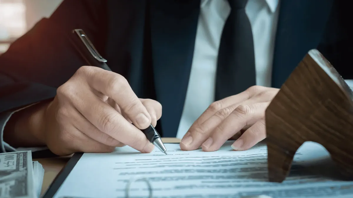 Close-up of hands signing a contract agreement next to a wooden house model and a stack of cash