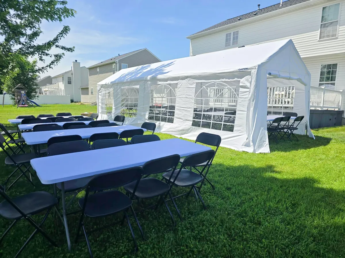 White party rental tent with window sidewalls, rectangular tables, and black folding chairs set up on a grassy lawn.