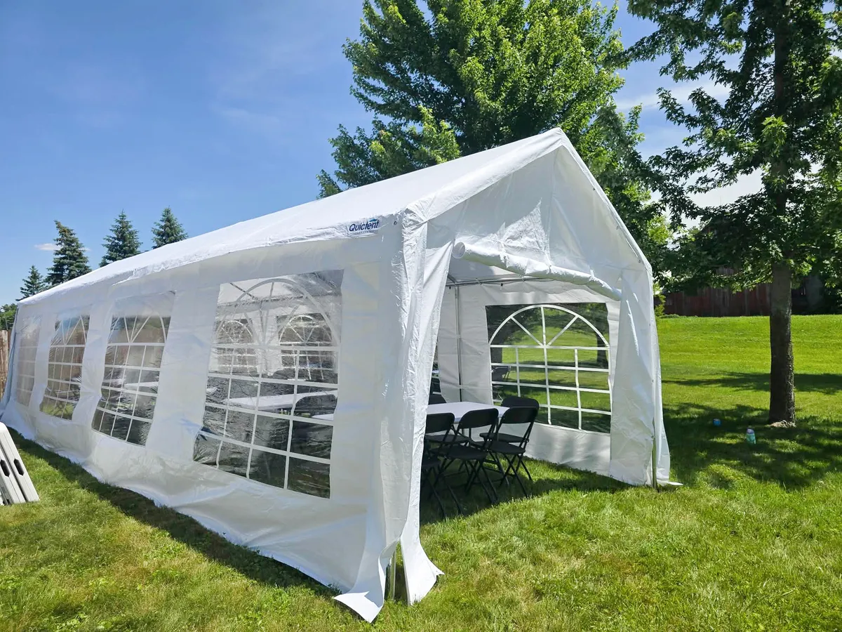 White party rental tent with window sidewalls, rectangular tables, and black folding chairs set up on a grassy lawn.