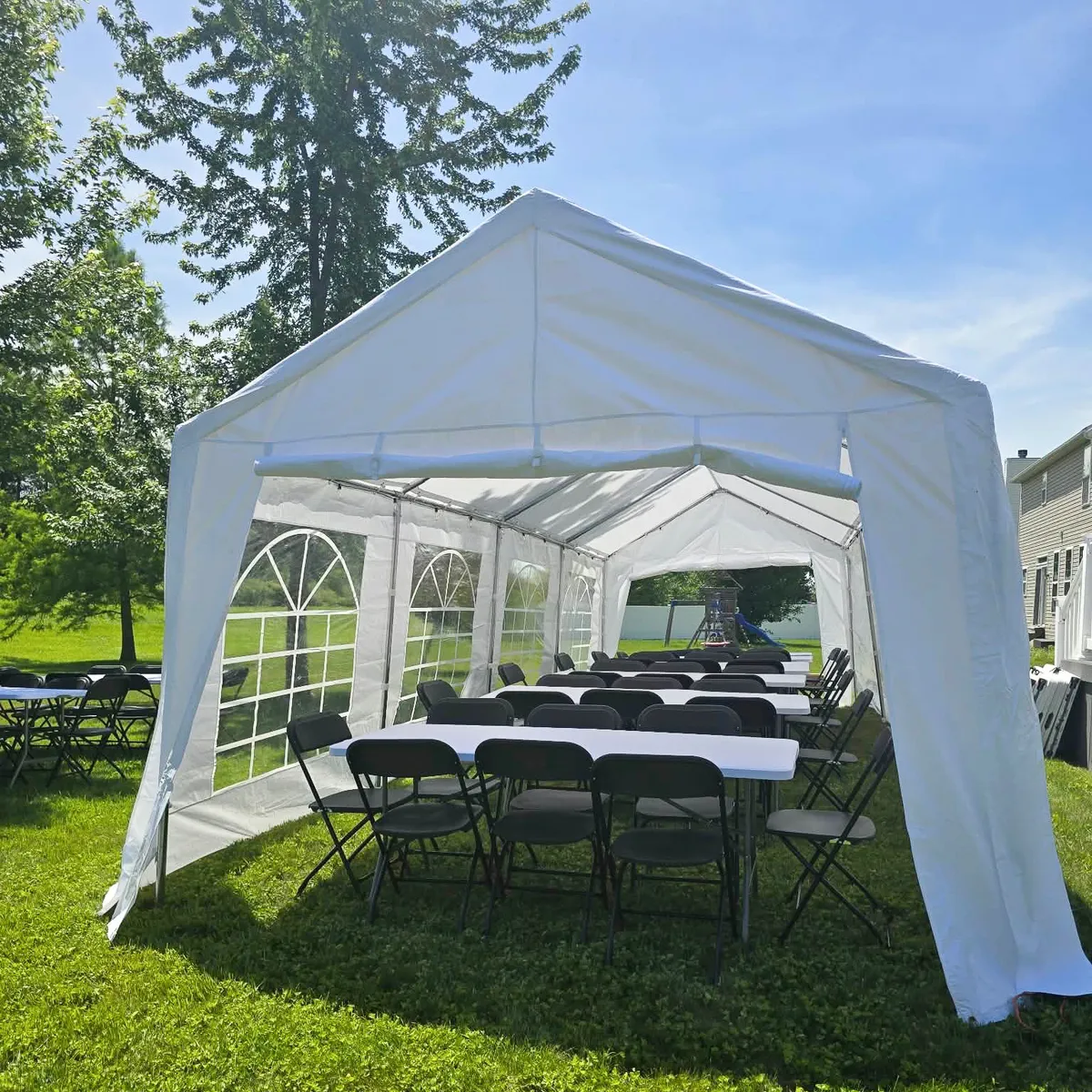 White party rental tent with window sidewalls, rectangular tables, and black folding chairs set up on a grassy lawn.