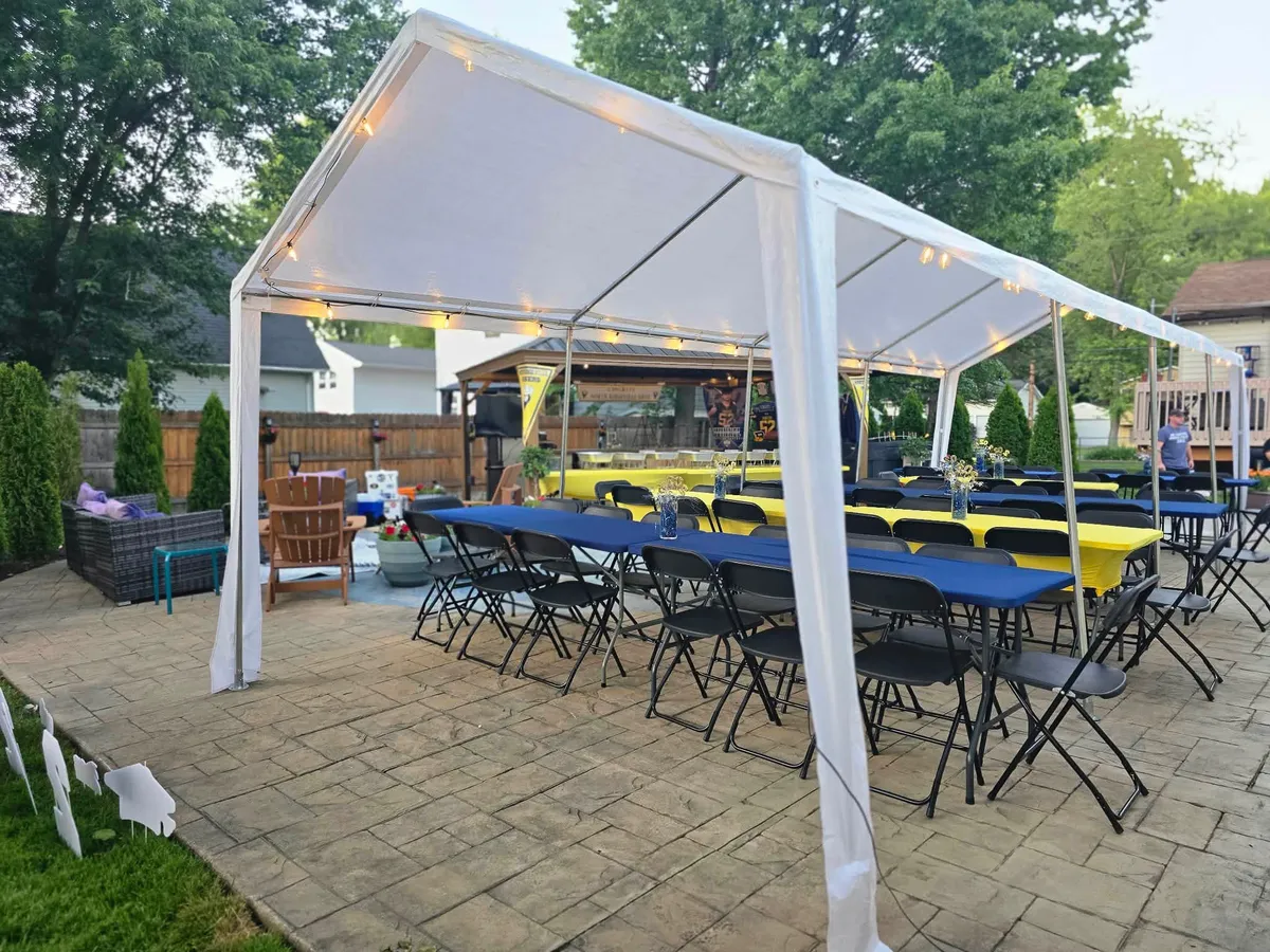 White party rental tent with window sidewalls, rectangular tables, and black folding chairs set up on a grassy lawn.