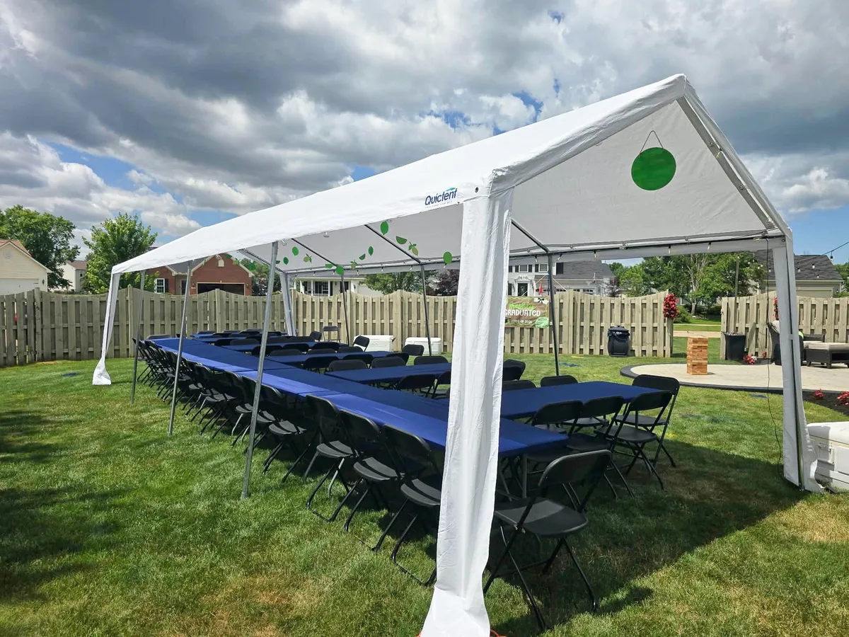 White party rental tent with window sidewalls, rectangular tables, and black folding chairs set up on a grassy lawn.