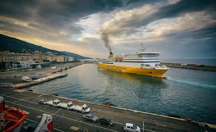 Un grand ferry jaune quitte un port côtier, de la fumée s'échappant de sa cheminée. Des voitures sont garées le long de la jetée, avec des montagnes visibles en arrière-plan sous un ciel nuageux.