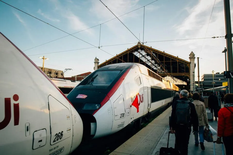 Un train TGV InOui en gare, prêt à partir, avec des passagers marchant sur le quai. La façade de la gare est visible à l'arrière-plan sous un ciel légèrement nuageux au coucher du soleil.