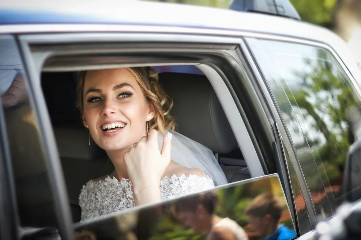 Une jeune mariée souriante, portant une robe de mariage ornée de dentelle, regarde par la fenêtre ouverte d'une voiture élégante, reflétant une atmosphère joyeuse et festive.