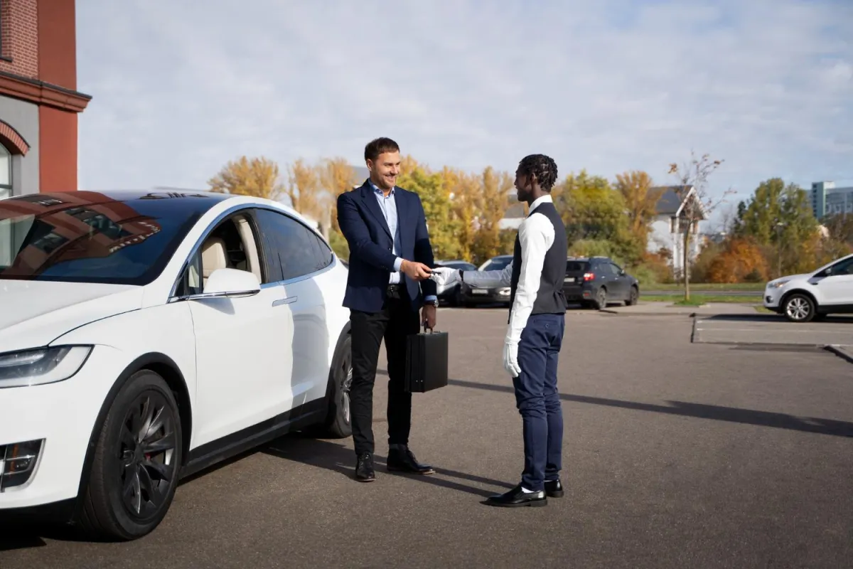 Chauffeur VTC professionnel en uniforme accueillant un client en costume près d’une voiture de luxe blanche sur un parking, idéal pour un transfert d’affaires ou un trajet haut de gamme. Décor automnal en arrière-plan.