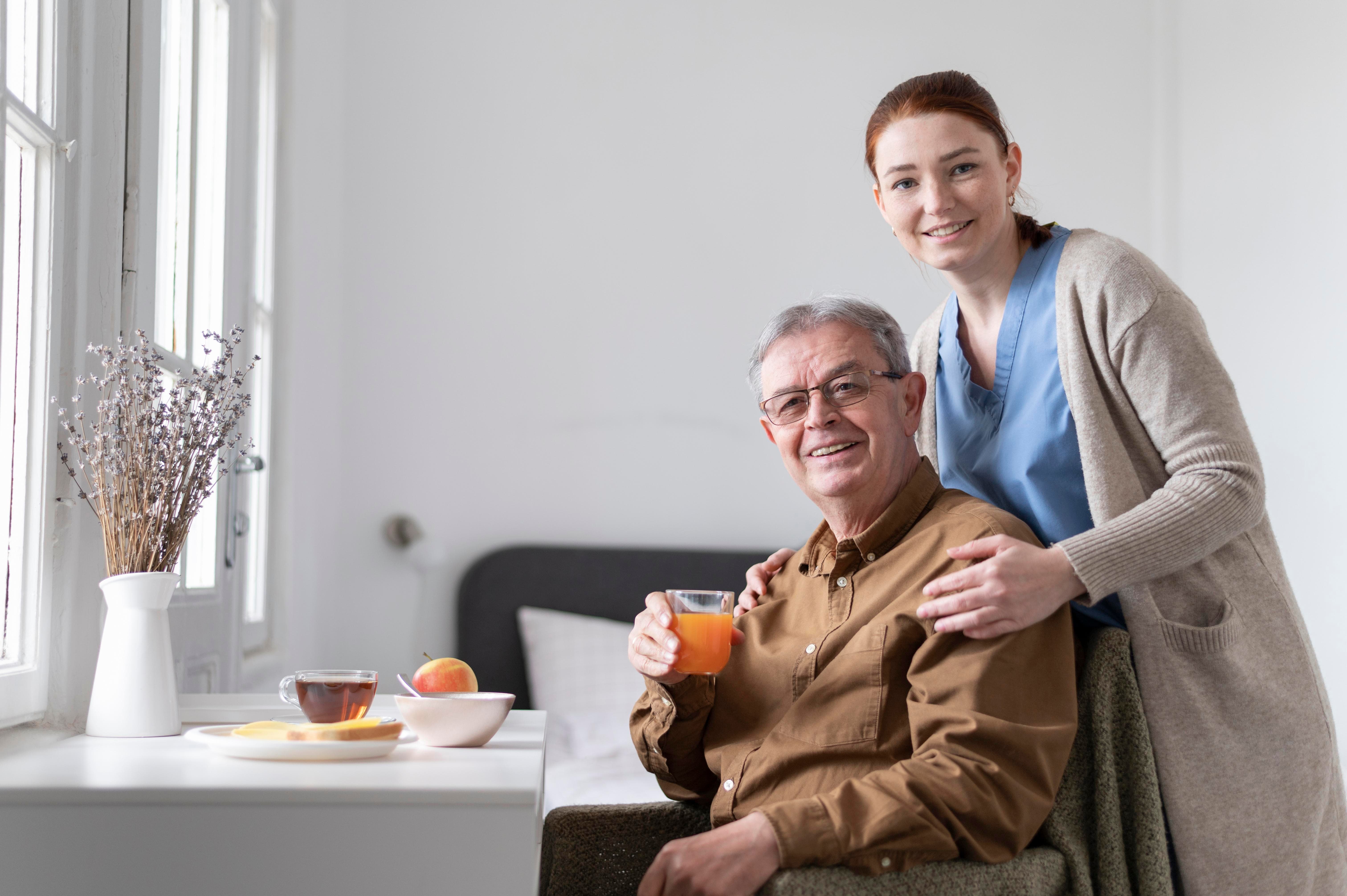 Smiling caregiver standing beside an elderly man seated with a glass of orange juice in a bright assisted living room, symbolising companionship and support.