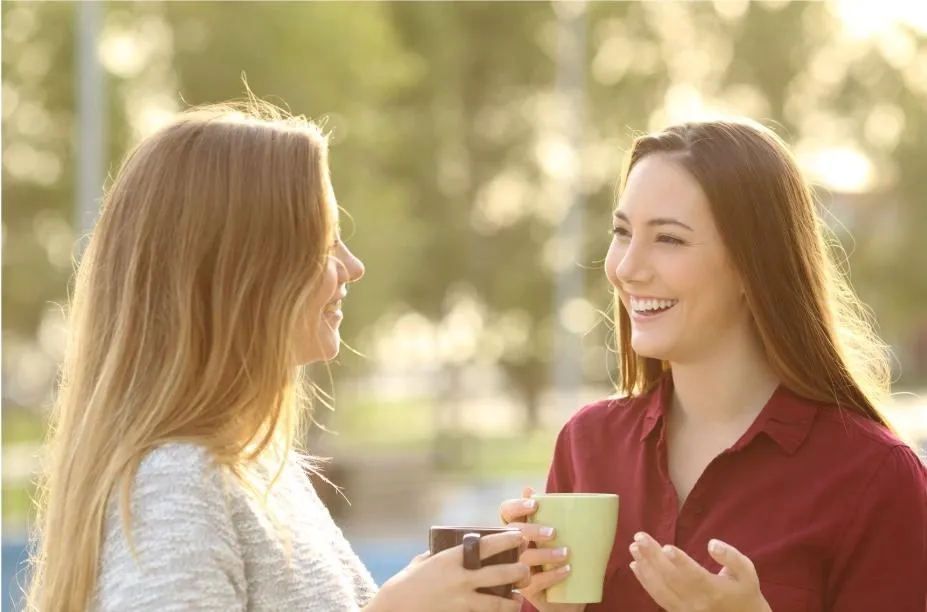 Two women smiling and holding coffee mugs while chatting outdoors.